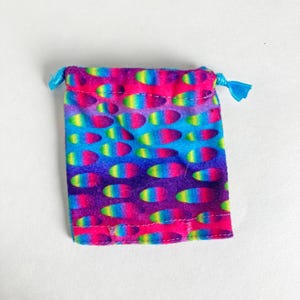 Puede incluir: Una pequeña bolsa rectangular con cordón y un patrón vibrante y colorido. La tela presenta un diseño repetido de formas ovaladas en tonos de rosa, azul, verde y amarillo sobre un fondo púrpura. El cordón es de color azul brillante.