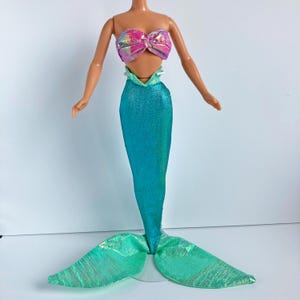 May include: A doll with a light skin tone featuring a strapless, iridescent pink and purple metallic top. The lower portion is a long, shimmering teal mermaid tail with a textured fabric finish that flares out into a wide, light green fin.