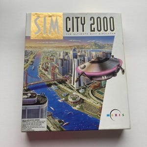 May include: Sim City 2000 video game box. The box features a cityscape with a bridge, river, and buildings. A futuristic flying saucer hovers above the city. The game title is in gold lettering.