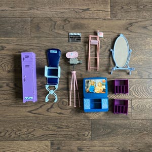 May include: A set of purple, blue, and pink plastic dollhouse furniture. The set includes a locker, a chair, a camera on a tripod, a television, a mirror, a ladder, and two small shelves.