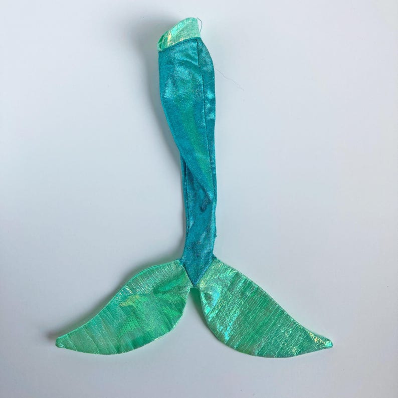 May include: A mermaid tail costume accessory crafted from a shimmering, iridescent turquoise and light green fabric. The piece features a long, slender body section that transitions into a wide, bifurcated fin at the base with a metallic sheen.