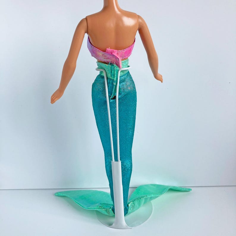 May include: A mermaid doll viewed from the back, featuring a pink and purple iridescent strapless top and a shimmering teal fabric tail. The doll is supported by a white plastic stand that holds the waist and extends down to the base.