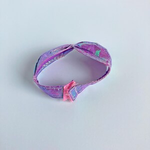 May include: A circular headband featuring a metallic purple and iridescent finish with a textured, ribbed surface. The band is joined at the back with a small, pink fabric hook-and-loop fastener, creating a continuous loop design.