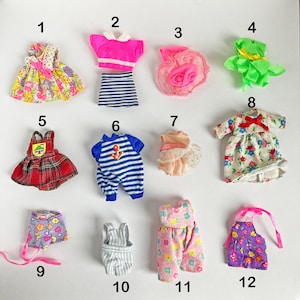 May include: A selection of twelve miniature doll outfits. The outfits include dresses, tops, skirts, and dungarees in various colours and patterns. Some outfits feature floral prints, stripes, and bows. The collection is suitable for doll enthusiasts.