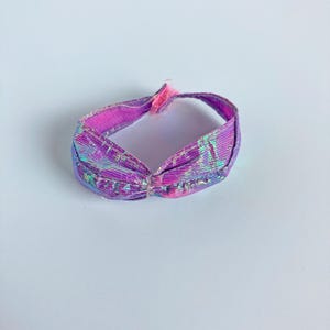 May include: A small, circular doll-sized headband featuring a vibrant, iridescent purple and pink metallic finish. The accessory has a gathered, ruched design at the front center and includes a small pink fabric tab closure at the back.