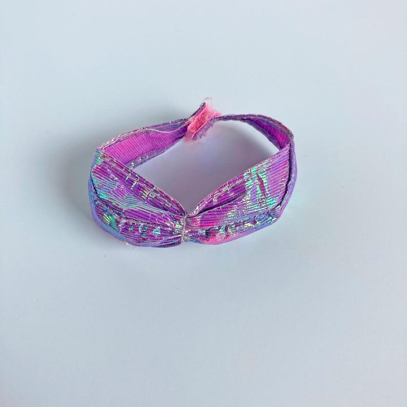 May include: A small, circular doll-sized headband featuring a vibrant, iridescent purple and pink metallic finish. The accessory has a gathered, ruched design at the front center and includes a small pink fabric tab closure at the back.