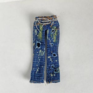 May include: Blue denim jeans with distressed details, featuring multiple rips and tears. The jeans have a decorative beaded belt and paint splatters. The jeans are a small size.