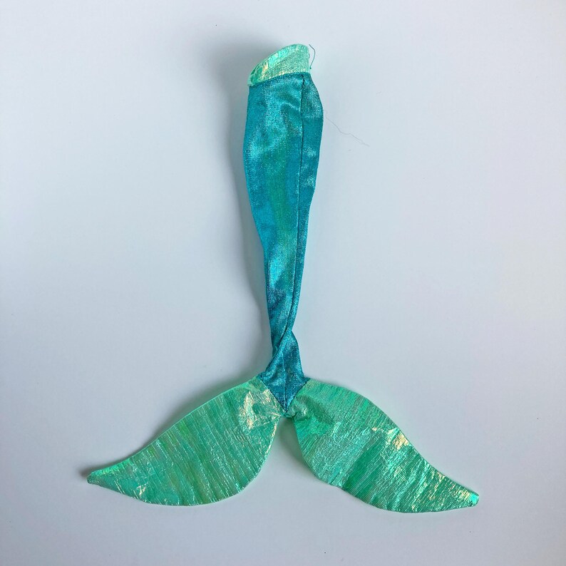 May include: A mermaid tail costume piece crafted from shimmering fabric in shades of teal and light green. The garment features a long, slender body section that transitions into a wide, bifurcated fin at the base with a metallic, iridescent finish.