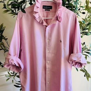 May include: A light pink long-sleeve button-down shirt featuring a ruffled collar, a front button placket, and a small embroidered logo on the chest. The shirt has rolled-up sleeves and a label inside the collar that reads Ralph Lauren.