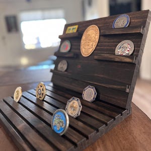 May include: A dark brown wooden display stand with slots for holding challenge coins. The stand has a slanted top with a shelf for displaying larger coins or patches. The stand is decorated with a dark wood stain and has a natural wood grain.