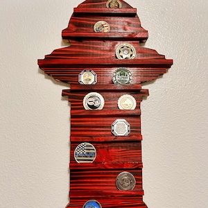 May include: A red-stained wooden display shaped like a fire hydrant, designed to hold challenge coins. The display has multiple shelves holding various coins with different designs and colors. The wood grain is visible, adding texture.