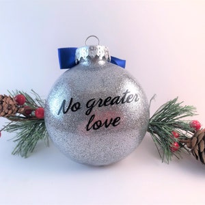 K&ouml;nnte beinhalten: Ein silberner Glitzer-Weihnachtsbaumschmuck mit einem blauen Band und dem Text "No greater love" in Schwarz.