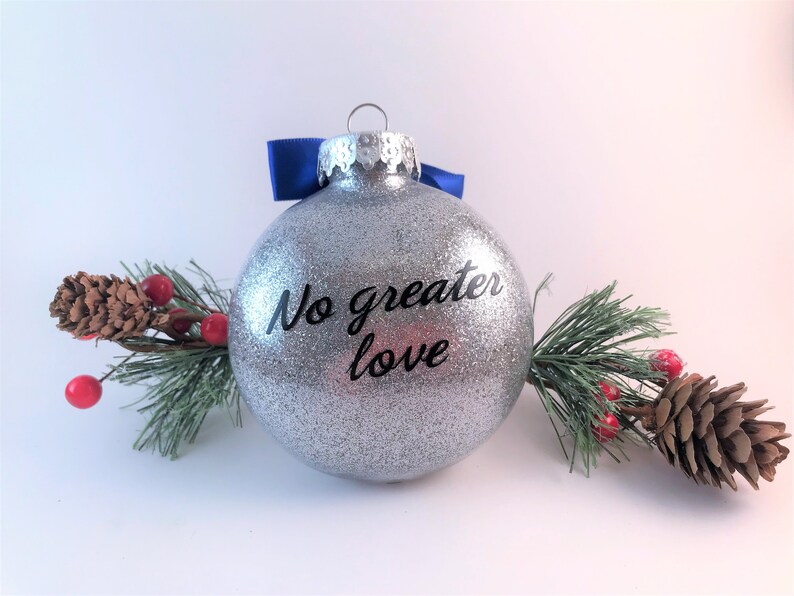 K&ouml;nnte beinhalten: Ein silberner Glitzer-Weihnachtsbaumschmuck mit einem blauen Band und dem Text "No greater love" in Schwarz.