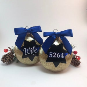 K&ouml;nnte beinhalten: Zwei goldene Weihnachtskugeln mit Glitzer und blauen Schleifen. Eine Kugel hat einen schwarzen Stern mit dem Wort "Wife" darauf. Die andere Kugel hat einen schwarzen Stern mit der Nummer "5264" darauf.