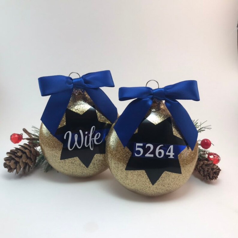 K&ouml;nnte beinhalten: Zwei goldene Weihnachtskugeln mit Glitzer und blauen Schleifen. Eine Kugel hat einen schwarzen Stern mit dem Wort "Wife" darauf. Die andere Kugel hat einen schwarzen Stern mit der Nummer "5264" darauf.