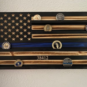 May include: Wooden American flag display with a blue line, featuring stars and stripes. The flag has shelves holding various challenge coins. The number "38412" is displayed on the flag. The flag is mounted on a white wall.