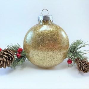 K&ouml;nnte beinhalten: Ein goldener Glitzer-Weihnachtsbaumschmuck mit silberner Spitze, umgeben von Tannenzweigen und roten Beeren.