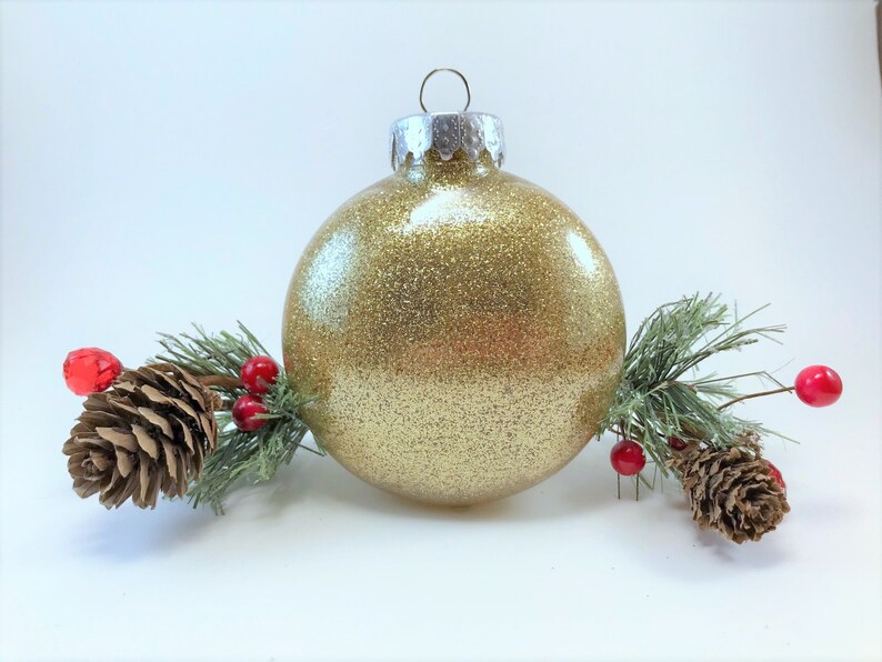 K&ouml;nnte beinhalten: Ein goldener Glitzer-Weihnachtsbaumschmuck mit silberner Spitze, umgeben von Tannenzweigen und roten Beeren.