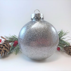 K&ouml;nnte beinhalten: Ein silberner Glitzer-Weihnachtsbaumschmuck mit silberner Kappe und &Ouml;se. Der Schmuck ist von Tannenzweigen mit roten Beeren umgeben.