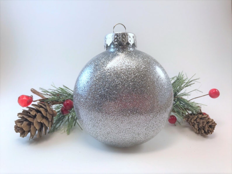 K&ouml;nnte beinhalten: Ein silberner Glitzer-Weihnachtsbaumschmuck mit silberner Kappe und &Ouml;se. Der Schmuck ist von Tannenzweigen mit roten Beeren umgeben.