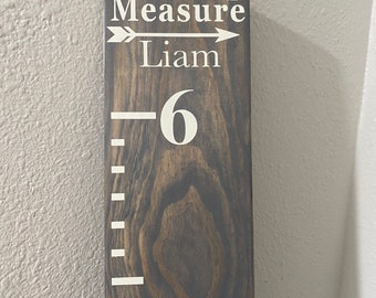 May include: A dark stained wooden growth chart ruler featuring white lettering that reads Loved Beyond Measure and the name Liam. The board includes a large number 6 and white horizontal tick marks for height measurement in inches.