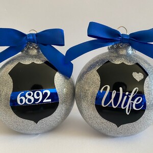 K&ouml;nnte beinhalten: Zwei silberne Glitzer-Weihnachtskugeln mit blauen Schleifen. Eine Kugel hat ein schwarzes Polizeibadge mit einem blauen Band und der Nummer "6892". Die andere Kugel hat ein schwarzes Polizeibadge mit einem blauen Band und dem Wort "Wife".