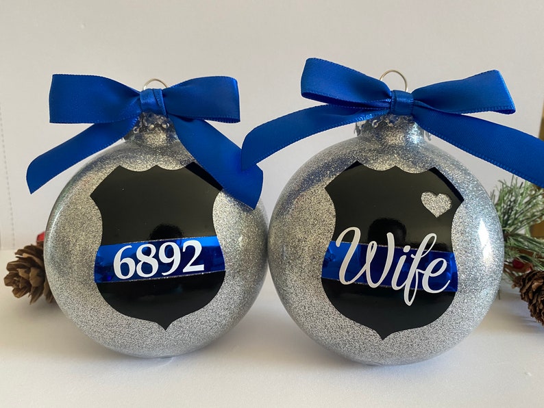 K&ouml;nnte beinhalten: Zwei silberne Glitzer-Weihnachtskugeln mit blauen Schleifen. Eine Kugel hat ein schwarzes Polizeibadge mit einem blauen Band und der Nummer "6892". Die andere Kugel hat ein schwarzes Polizeibadge mit einem blauen Band und dem Wort "Wife".