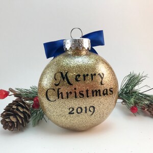 K&ouml;nnte beinhalten: Goldene Weihnachtskugel mit Glitzer, blauem Band und dem Text "Frohe Weihnachten 2019".