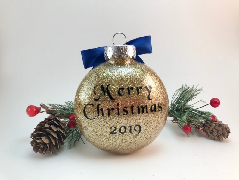 K&ouml;nnte beinhalten: Goldene Weihnachtskugel mit Glitzer, blauem Band und dem Text "Frohe Weihnachten 2019".