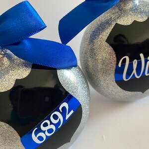 K&ouml;nnte beinhalten: Zwei silberne Glitzer-Weihnachtskugeln mit einem schwarzen und blauen Polizeibadge-Design. Eine Kugel tr&auml;gt das Wort "Wife" und die andere die Nummer "6892".