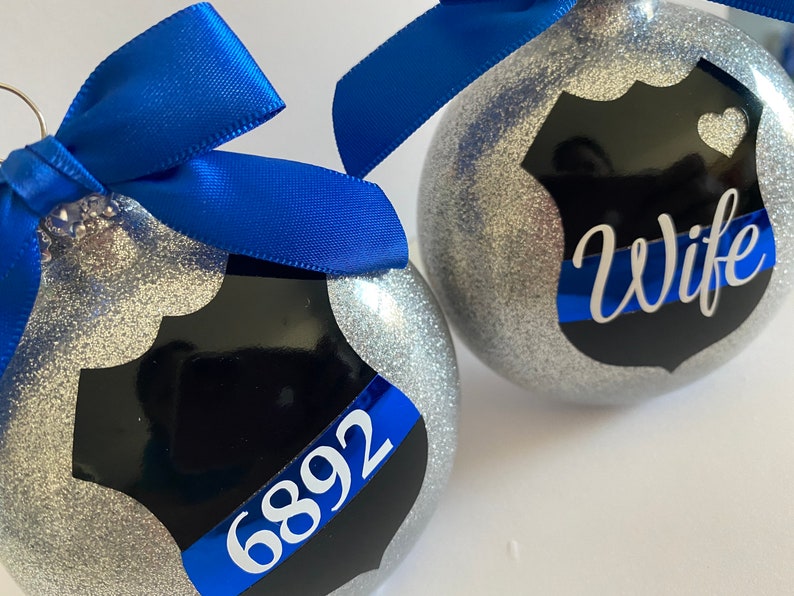 K&ouml;nnte beinhalten: Zwei silberne Glitzer-Weihnachtskugeln mit einem schwarzen und blauen Polizeibadge-Design. Eine Kugel tr&auml;gt das Wort "Wife" und die andere die Nummer "6892".