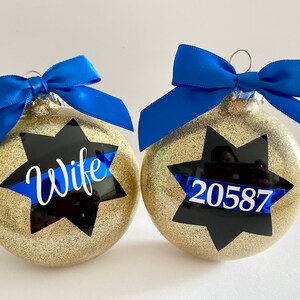 K&ouml;nnte beinhalten: Zwei goldene Weihnachtskugeln mit Glitzer und blauen B&auml;ndern. Eine Kugel hat einen schwarzen Stern mit einem blauen Band und dem Wort "Wife" darauf. Die andere Kugel hat einen schwarzen Stern mit einem blauen Band und der Nummer "20587" darauf.