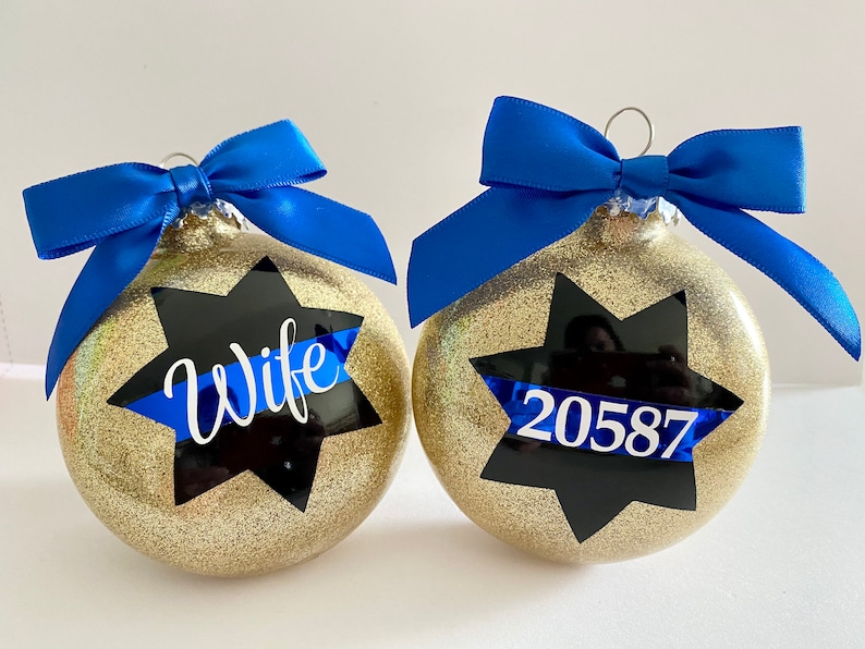 K&ouml;nnte beinhalten: Zwei goldene Weihnachtskugeln mit Glitzer und blauen B&auml;ndern. Eine Kugel hat einen schwarzen Stern mit einem blauen Band und dem Wort "Wife" darauf. Die andere Kugel hat einen schwarzen Stern mit einem blauen Band und der Nummer "20587" darauf.