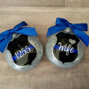 K&ouml;nnte beinhalten: Zwei silberne Glitzer-Weihnachtskugeln mit einem schwarzen und blauen Polizeibadge-Design. Eine Kugel sagt "Wife" und die andere sagt "1255".