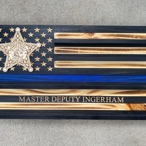 May include: Wooden American flag with a blue line and a sheriff's badge. The flag features the text "MASTER DEPUTY INGERHAM" and a star-shaped badge with the words "DEPUTY SHERIFF ORANGE COUNTY FLORIDA".