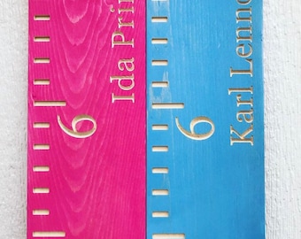 May include: Two wooden growth charts, one hot pink and one blue. Each chart has a carved number 6 and a name. The pink chart says "Ida Prine" and the blue chart says "Karl Lennox". Both charts have measurement markings.