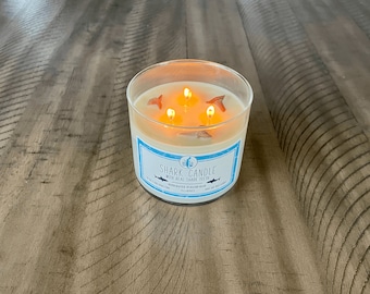 May include: A clear glass candle jar with a white candle and three lit wicks. The candle is decorated with real shark teeth and the label reads "Shark Candle with Real Shark Teeth".