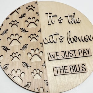 May include: A wooden sign with a paw print and fishbone pattern on one half and the text "It's the cat's house we just pay the bills" on the other half.