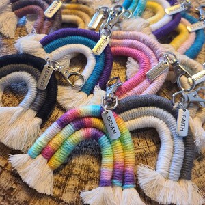 May include: A collection of colorful macrame rainbow keychains. Each keychain features a rainbow design with a silver clasp and a small metal tag with words like "Dream", "Hope", and "Faith". The keychains are made of various colored threads.
