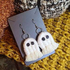 May include: A pair of white crocheted ghost earrings with black glitter eyes hanging on a gray card. The earrings are on a background of yellow and white knitted fabric.