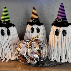 May include: Three white ghost-shaped decorations with black and white striped witch hats. The hats have black bows and black buttons. The ghosts are made of white yarn and have black eyes. A sparkly gold and brown pumpkin sits in front of the ghosts.