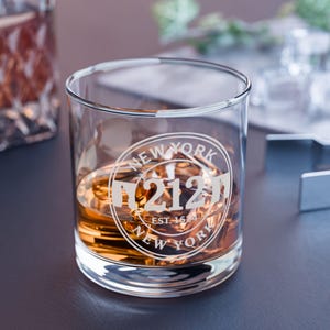 Puede incluir: Un vaso de whisky redondo y transparente lleno de líquido ámbar. El vaso presenta un diseño circular blanco con las palabras "NEW YORK" y "212" impresas. El vaso está sobre una superficie oscura.