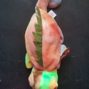 May include: A tie-dye green, pink, and orange chameleon beanie baby with a green felt spine. The beanie baby has a red tag with the words "Ty" and "Beanie Babies" on it.