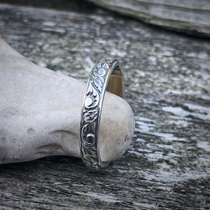 May include: A silver band ring with an intricate floral design. The ring is made of polished metal and has a smooth, rounded surface.
