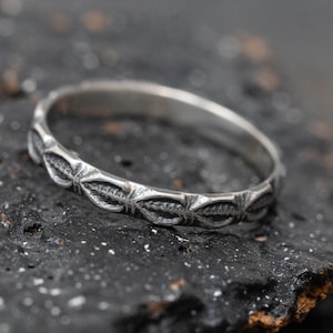 Puede incluir: Un anillo de banda de plata con un patrón decorativo de formas de hojas repetidas.