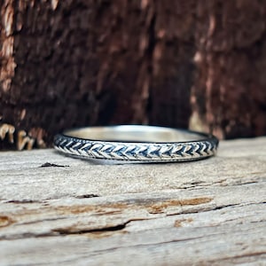 Könnte beinhalten: Ein silberner Ring mit einem detaillierten Fischgrätenmuster. Der Ring liegt auf einer rustikalen Holzoberfläche, mit einem verschwommenen Hintergrund aus dunkelbraunem Holz. Das Design des Rings ist eine sich wiederholende Reihe von V-Formen.