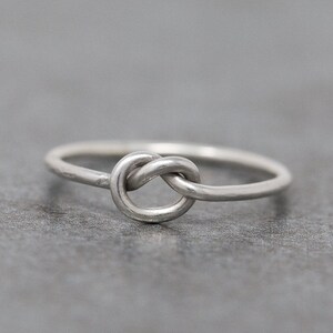 May include: A silver-colored ring featuring a knot design. The ring is simple and elegant, with a smooth, polished finish. The knot is the focal point, adding a unique and symbolic touch to the jewelry. The ring is shown on a gray surface.