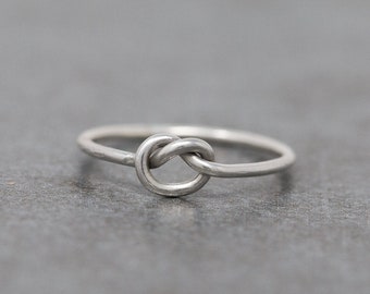Op de afbeelding: Een zilverkleurige ring met een knoopontwerp. De ring heeft een eenvoudig, elegant ontwerp met een gladde, gepolijste afwerking. De knoop is het centrale kenmerk en geeft het sieraad een unieke en symbolische uitstraling. De ring wordt getoond op een grijze ondergrond.