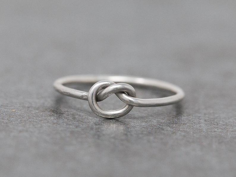 May include: A silver-colored ring featuring a knot design. The ring is simple and elegant, with a smooth, polished finish. The knot is the focal point, adding a unique and symbolic touch to the jewelry. The ring is shown on a gray surface.