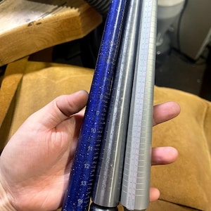 May include: Three metal ring sizers, one blue, one silver, and one gray, with size markings in inches and millimeters. The sizers are held in a hand.